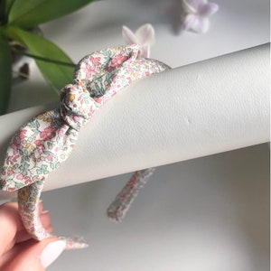 May include: A fabric-covered headband featuring a central knotted detail. The material has a light-colored background with a dense, multi-colored floral pattern in shades of pink, green, and yellow, creating a delicate and intricate botanical design.