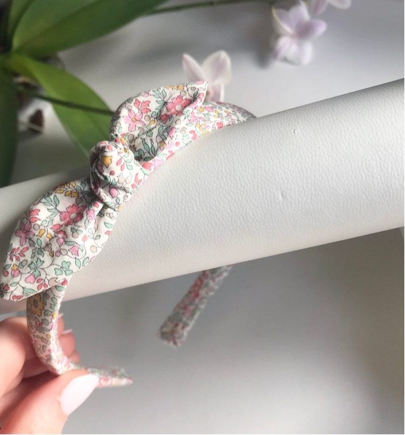 May include: A fabric-covered headband featuring a central knotted detail. The material has a light-colored background with a dense, multi-colored floral pattern in shades of pink, green, and yellow, creating a delicate and intricate botanical design.