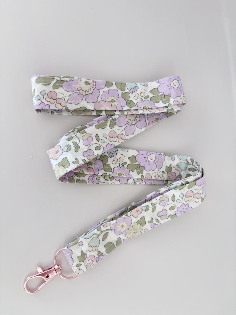 May include: A floral lanyard in a pattern of light purple, pink, and green. It features a rose gold-coloured clasp. The fabric is a light, delicate material.