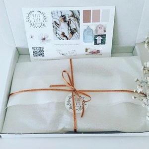 May include: A white gift box with a brown ribbon and a small round tag with the text "Wild Ivy" on it. The box is filled with white tissue paper and is sitting on a white surface. There is a bouquet of white flowers in the background.