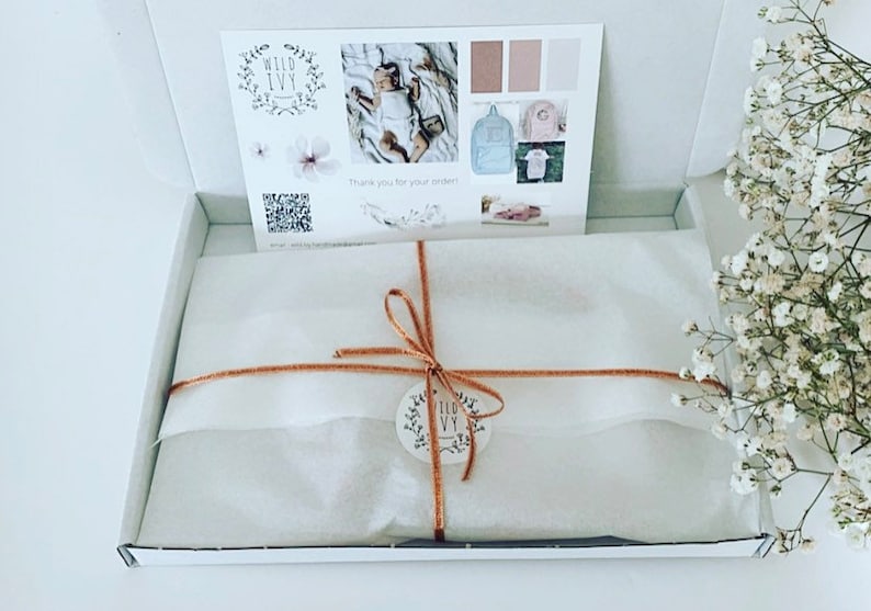 May include: A white gift box with a brown ribbon and a small round tag with the text "Wild Ivy" on it. The box is filled with white tissue paper and is sitting on a white surface. There is a bouquet of white flowers in the background.