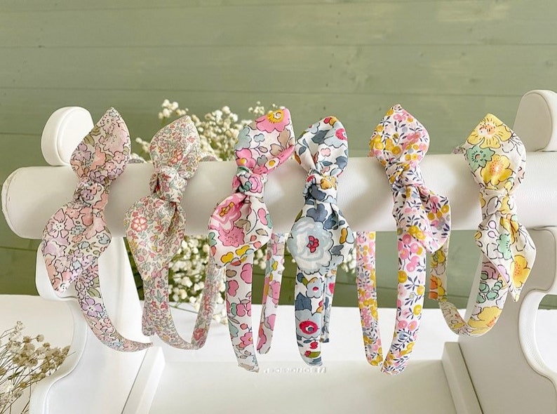 May include: Six fabric-covered headbands arranged on a white display stand. Each headband features a unique floral pattern in various colors including pink, yellow, blue, and green, and is topped with a decorative knotted bow detail at the center.