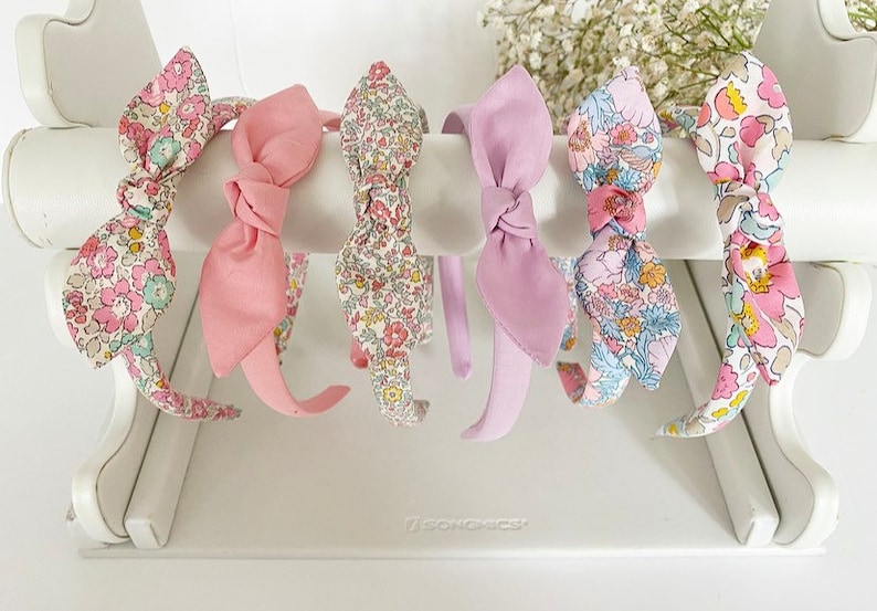 May include: Six headbands with floral patterns and bows. The headbands are displayed on a white stand. The headbands are in shades of pink, white, and green.