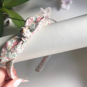 May include: A white headband with a floral print bow. The bow is tied in a knot and the headband is made of a soft fabric.