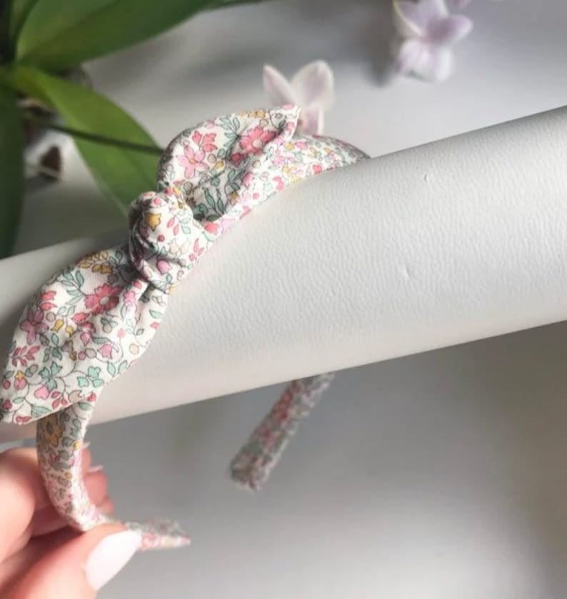 May include: A white headband with a floral print bow. The bow is tied in a knot and the headband is made of a soft fabric.