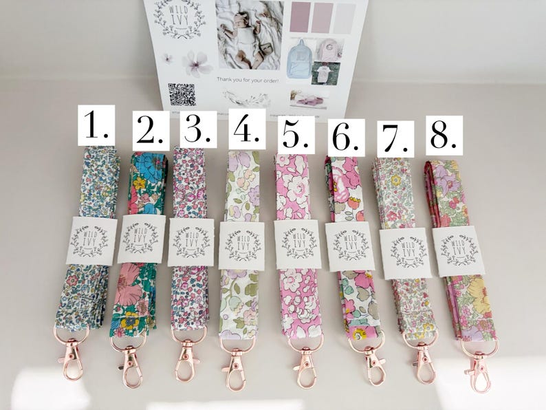 May include: Eight floral lanyards with rose gold clasps. Each lanyard displays a different colourful floral pattern, with a white tag that reads "Wild Ivy". The lanyards are arranged on a white surface.