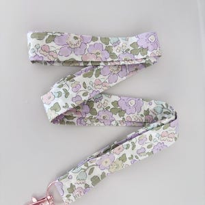 May include: A floral patterned lanyard in shades of lilac, pink, and green on a white base. It features a rose gold-coloured clasp.