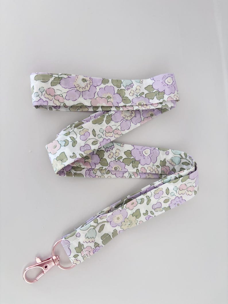 May include: A floral patterned lanyard in shades of lilac, pink, and green on a white base. It features a rose gold-coloured clasp.