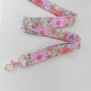 May include: A floral patterned lanyard with a rose gold-coloured clasp. The fabric displays pink, purple, and yellow flowers on a light green background. The lanyard is designed to be worn around the neck.