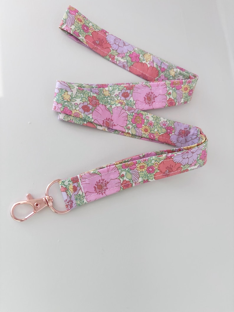 May include: A floral patterned lanyard with a rose gold-coloured clasp. The fabric displays pink, purple, and yellow flowers on a light green background. The lanyard is designed to be worn around the neck.
