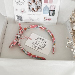 May include: A pink and white floral headband with a bow on the side. The headband is packaged in a white box with a small tag that says "Wild Ivy Handmade".