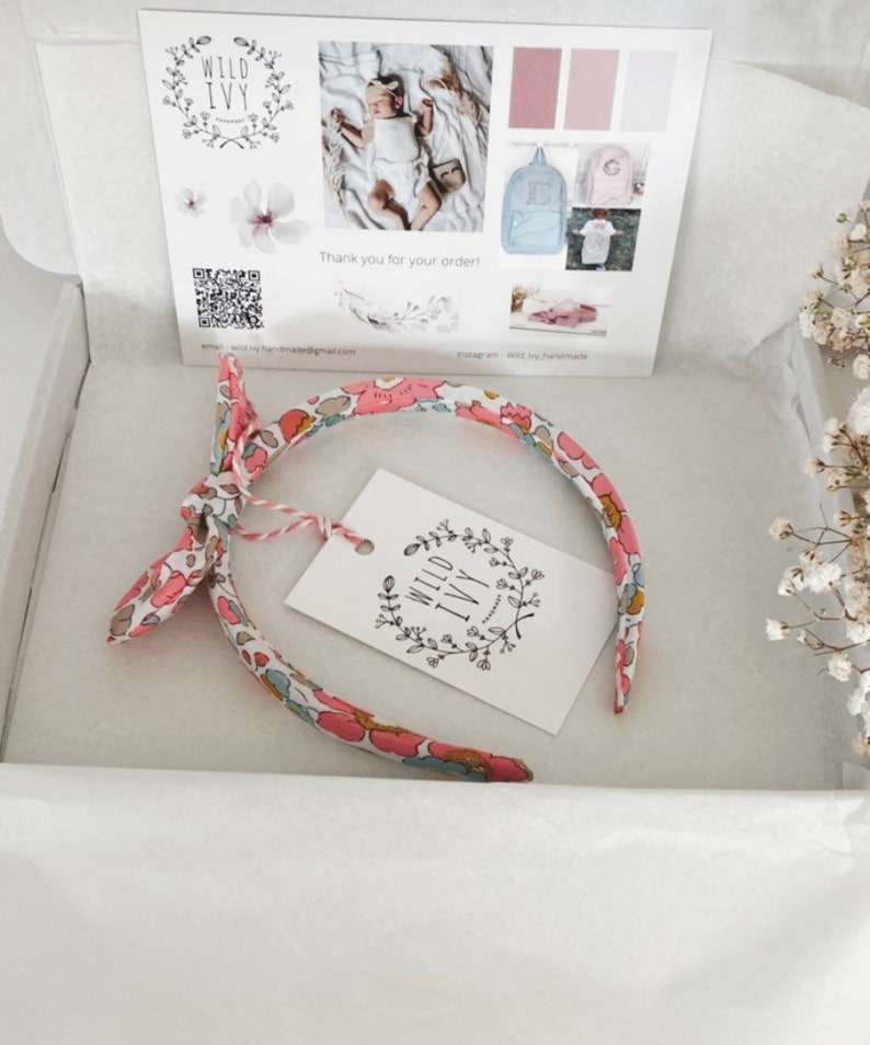 May include: A pink and white floral headband with a bow on the side. The headband is packaged in a white box with a small tag that says "Wild Ivy Handmade".