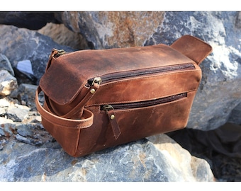 May include: A brown leather travel toiletry bag with two zippered compartments and a top handle. The bag is sitting on a pile of rocks.
