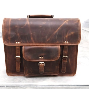 May include: A brown leather satchel bag with a front flap and a large front pocket. The bag has two buckles on each side and a metal buckle on the front pocket.