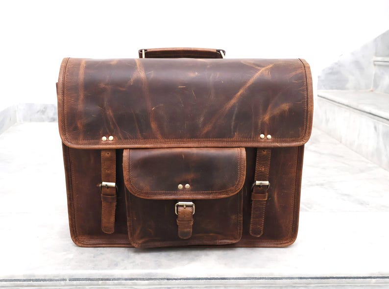 May include: A brown leather satchel bag with a front flap and a large front pocket. The bag has two buckles on each side and a metal buckle on the front pocket.
