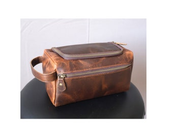 Puede incluir: Una bolsa de viaje de cuero marrón con cierre de cremallera y asa. La bolsa tiene un aspecto desgastado y está hecha de cuero de alta calidad.