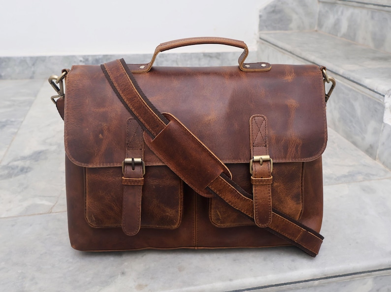 May include: A brown leather messenger bag with two front pockets and a long adjustable strap. The bag has a top handle and brass buckles.