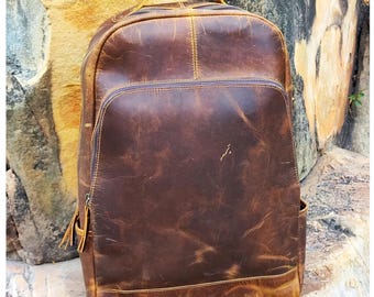 May include: A brown leather backpack with a distressed look. The backpack has a single large compartment with a zipper closure and a smaller pocket on the front. The backpack has a top handle and adjustable shoulder straps.
