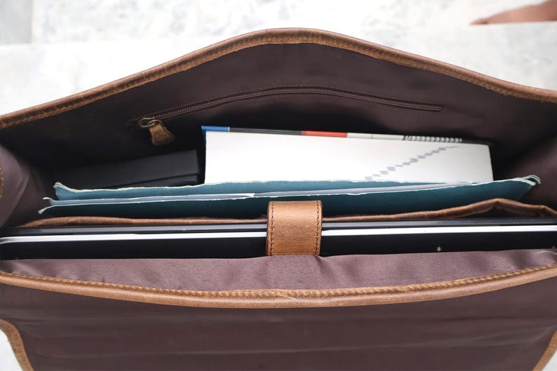 May include: A brown leather messenger bag with a zippered pocket and a flap closure. The bag is open and contains a laptop, a notebook, and other items.