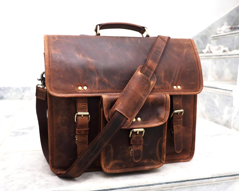 May include: A brown leather messenger bag with a large front pocket and adjustable shoulder strap. The bag has brass buckles and rivets.