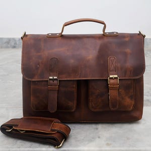 May include: A brown leather briefcase with two front pockets and a long adjustable shoulder strap. The bag has a top handle and brass buckles.