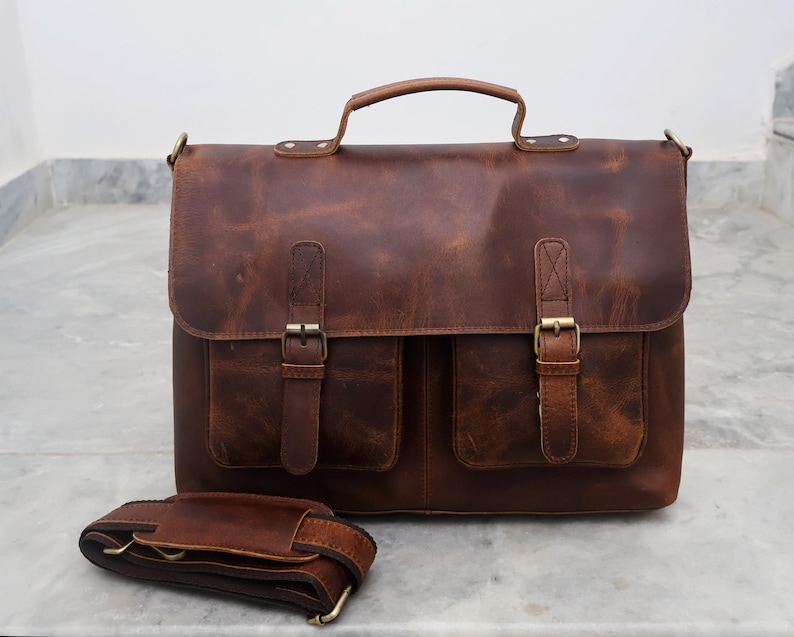 May include: A brown leather briefcase with two front pockets and a long adjustable shoulder strap. The bag has a top handle and brass buckles.