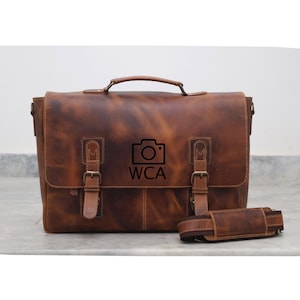 May include: A brown leather messenger bag with a camera logo and the text "WCA". The bag has two buckles on the front and a long adjustable strap.
