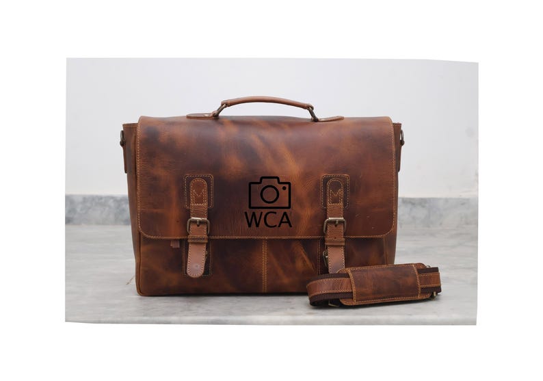 May include: A brown leather messenger bag with a camera logo and the text "WCA". The bag has two buckles on the front and a long adjustable strap.