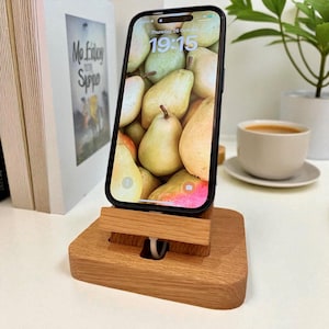 Può includere: Un supporto per telefono in legno con un telefono che mostra uno sfondo di pere. Il supporto è in legno marrone chiaro e ha una fessura per tenere il telefono. Un cavo bianco è visibile. Una tazza di caffè e una pianta sono sullo sfondo.