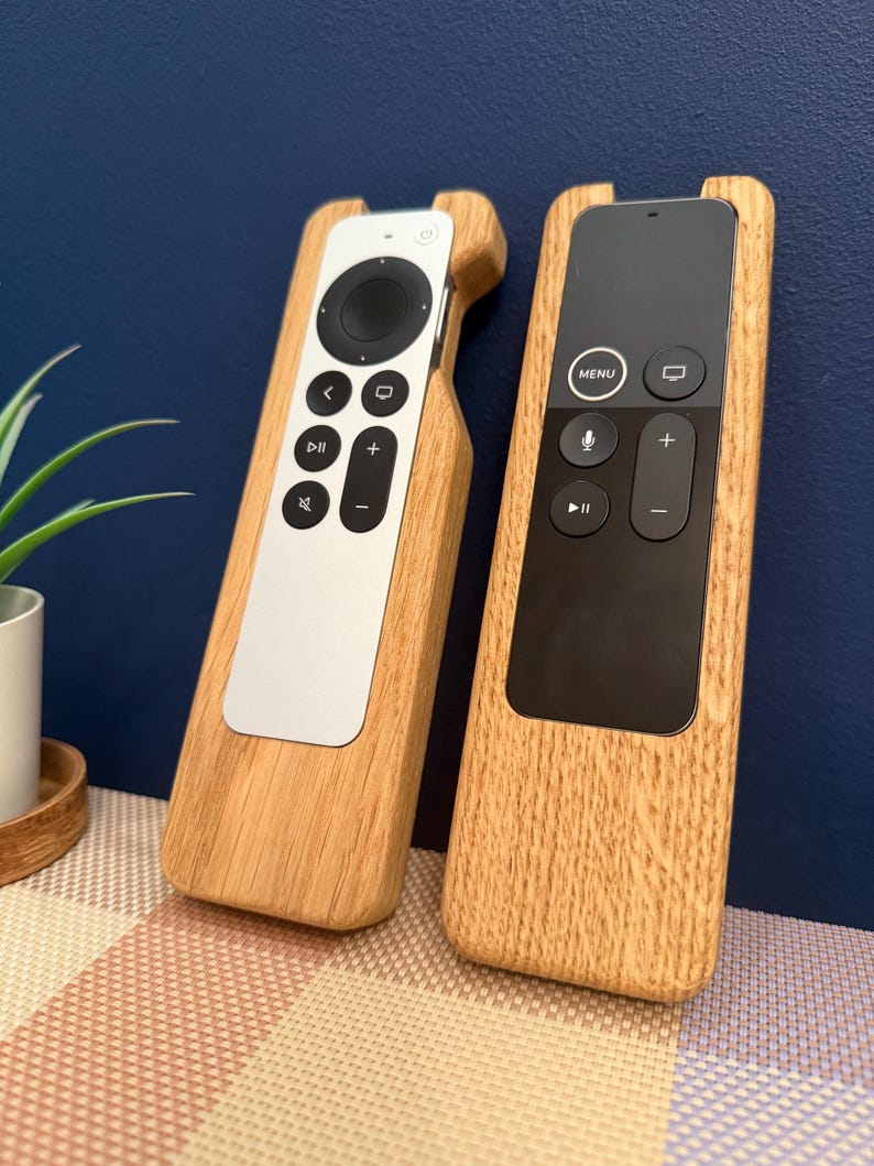 Oak case for Apple TV remote. smooth contoured corners, smooth finish. Remote sits flush into the holder and stays in position using micro suction pads. image shows both variants of the Apple TV remote on a desktop.