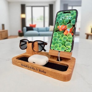 Op de afbeelding: Een houten bureau-organizer met een telefoon, bril en oortelefoons. De telefoon toont een kleurrijk scherm met vissen. De organizer heeft een sleuf voor de telefoon, een standaard voor brillen en een dienblad met de tekst "Brad's Bits & Bobs".