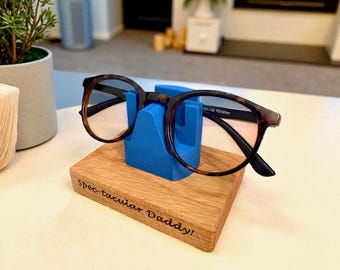 May include: Round-framed eyeglasses with a tortoiseshell pattern rest on a blue holder atop a wooden base. The base is engraved with the words "Spec-tacular Daddy!" The eyeglasses have clear lenses and black arms.