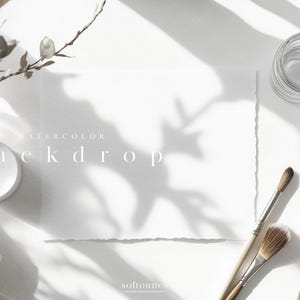 May include: A bright, overhead shot of a white watercolor backdrop with the text "watercolor backdrop". The scene includes paintbrushes, small glass jars, and a sprig of a plant. The scene is lit with natural light and shadows.