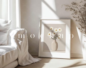 Op de afbeelding: Een witte ingelijste print met een beige achtergrond waarop een boeket witte madeliefjes in een beige vaas staat. De print hangt aan een witte muur in een kamer met een witte bank en een raam.