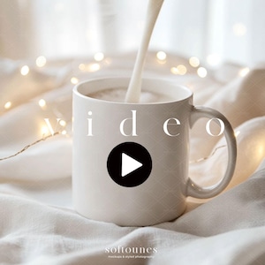 May include: A white ceramic mug with a handle is being filled with a creamy liquid. The word "video" is displayed on the mug, with a black play button icon. The background features soft, neutral tones and fairy lights.