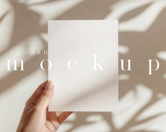 May include: A white A2 card mockup held by a hand, with the word "mockup" and "A2 CARD" printed on the card. The background is a neutral beige with shadows of leaves. The text "softounes" is printed at the bottom.