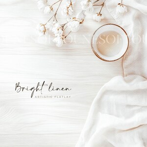 Puede incluir: Una tela de lino blanco drapeada sobre una superficie de madera blanca con flores blancas y un tazón blanco. El texto "Bright linen" y "Artistic Flatlay" está escrito en negro.
