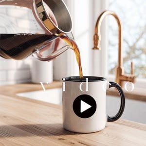 May include: Coffee being poured into a white mug with a black interior and handle. The mug has a black circle with a white play button. The word "video" is written on the mug. A gold faucet is in the background.
