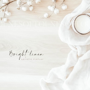 May include: A flat lay image of a white linen cloth draped over a light wood surface. A white ceramic mug filled with a white liquid sits on the cloth. White cotton flower stems are arranged in the upper left and right corners of the image. The text "Bright linen" and "Artistic Flatlay" is written in black script on the wood surface.