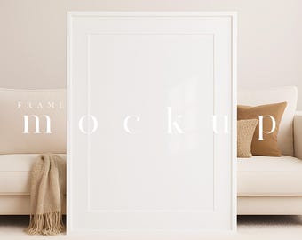 Op de afbeelding: Een grote witte fotolijst-mockup, leunend tegen een crèmekleurige bank met sierkussens en een deken met franjes. Het woord "mockup" wordt in witte tekst op het frame weergegeven. De achtergrond is een neutraal beige.