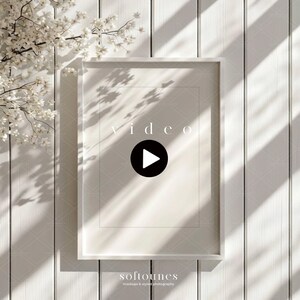 Pode incluir: Uma maquete de vídeo emoldurada em branco com a palavra "vídeo" em uma fonte clara. Um botão de reprodução preto está no centro. A moldura está contra um fundo de madeira branca com sombras e um galho floral.