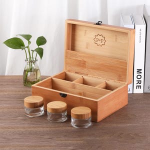 May include: A bamboo storage box with an open lid, revealing divided compartments. Three small glass jars with wooden lids are in front of the box. A small plant in a glass vase is to the left. The box has the text "P+P" inside a starburst.