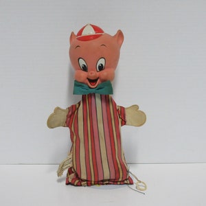 May include: Vintage Porky Pig hand puppet. The puppet features a pink pig head with a red and white hat, a green bow tie, and a striped shirt in red, pink, and beige. The puppet has beige hands and a string attached.