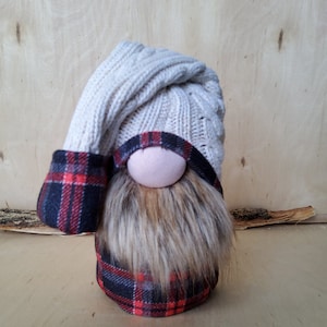May include: A gnome doll with a white knitted hat and a red and black plaid shirt. The gnome has a brown beard and a pink nose.