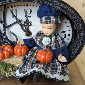 May include: A decorative clock with a black ornate frame, featuring a doll dressed in plaid clothing and a blue hat. The doll is surrounded by small orange pumpkins. The clock hands are black.