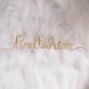 Peut inclure: Une œuvre d'art en fil d'or épelant "Brottelheim" dans une police cursive. Le mot est centré sur un fond blanc et duveteux. Le fil est fin et délicat, avec un petit bijou au milieu du mot.