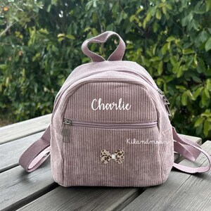 Peut inclure: Un sac à dos en velours côtelé rose poussiéreux avec le nom "Charlie" en écriture blanche. Il comporte une poche avant zippée, un petit nœud et des bretelles réglables. Le sac à dos a une poignée en forme de cœur. Le texte "Kikandmoon" est également visible.
