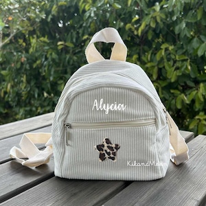 Peut inclure: Un sac à dos en velours côtelé gris clair avec une poignée et des bretelles crème. Le sac à dos comporte une poche avant zippée, le nom "Alycia" en écriture cursive et un écusson en forme d'étoile marron et blanc. La marque "KikandMom" est également visible.