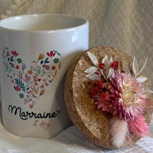 Peut inclure: Mug en céramique blanche avec un motif floral en forme de cœur et le texte "Marraine L'Ary". Un couvercle en liège est orné de fleurs séchées roses, rouges et blanches. Le mug et le couvercle sont posés sur un fond neutre.