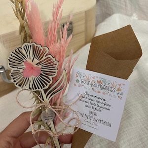 Op de afbeelding: Een boeket van gedroogde bloemen met een houten bloemaccent en een klein label. Het boeket bevat roze pampasgras en tarwe. Een kleine kaart in een bruine papieren kegel leest "BONNES VACANCES".